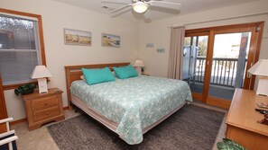 Quarto com cama, duas almofadas, mesa de cabeceira, abajur, janela com persianas, ventoinha no teto e vista para o exterior.