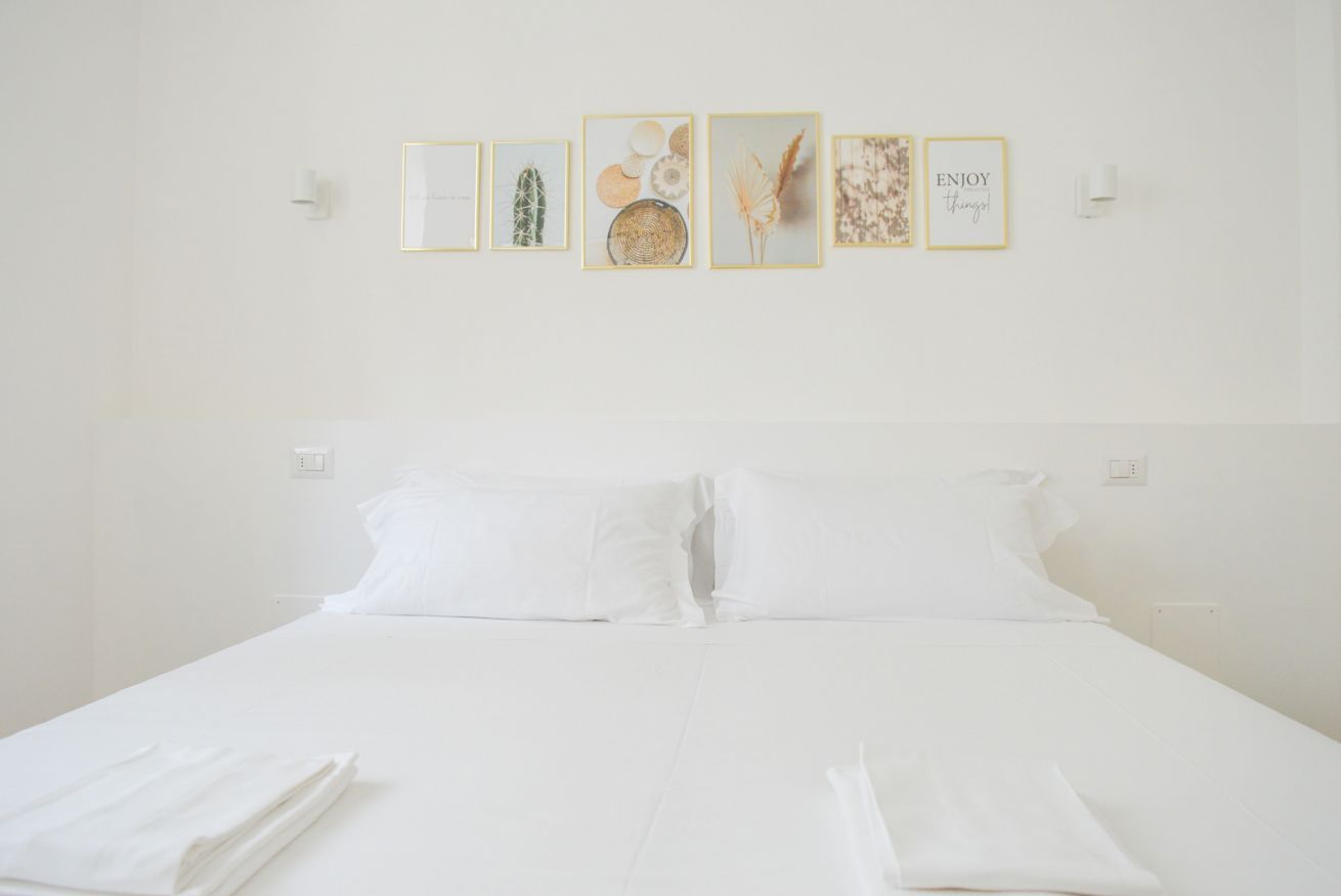 Un letto rifatto con lenzuola e cuscini bianchi, una parete con opere d'arte incorniciate e una parete bianca con interruttori.