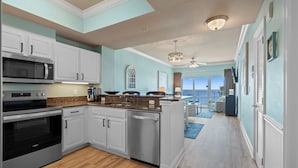 A kitchen with white cabinets, a stainless steel oven, and a dishwasher. There is a view of a balcony with a chair and a table. - Crystal Tower 908 | Gulf Shores Beachfront | 3 Pools, Hot Tub, Lazy River (Gulf Shores)