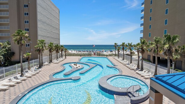 A swimming pool with geometric patterns, surrounded by palm trees and lounge chairs, with a view of the ocean and a hotel building in the background. - Crystal Tower 908 | Gulf Shores Beachfront | 3 Pools, Hot Tub, Lazy River (Gulf Shores)