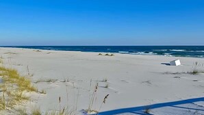 Ein Strand mit weißem Sand und spärlicher Vegetation, ein klarer, blauer Himmel und das Meer im Hintergrund.