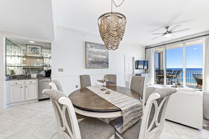 A dining area with a round table, chairs, a chandelier, and a view of the ocean through large windows. - Spanish Key 507 | Steps from Flora Bama | Oceanfront Condo | Pools, Hot Tub, Gym & More! (Pensacola)