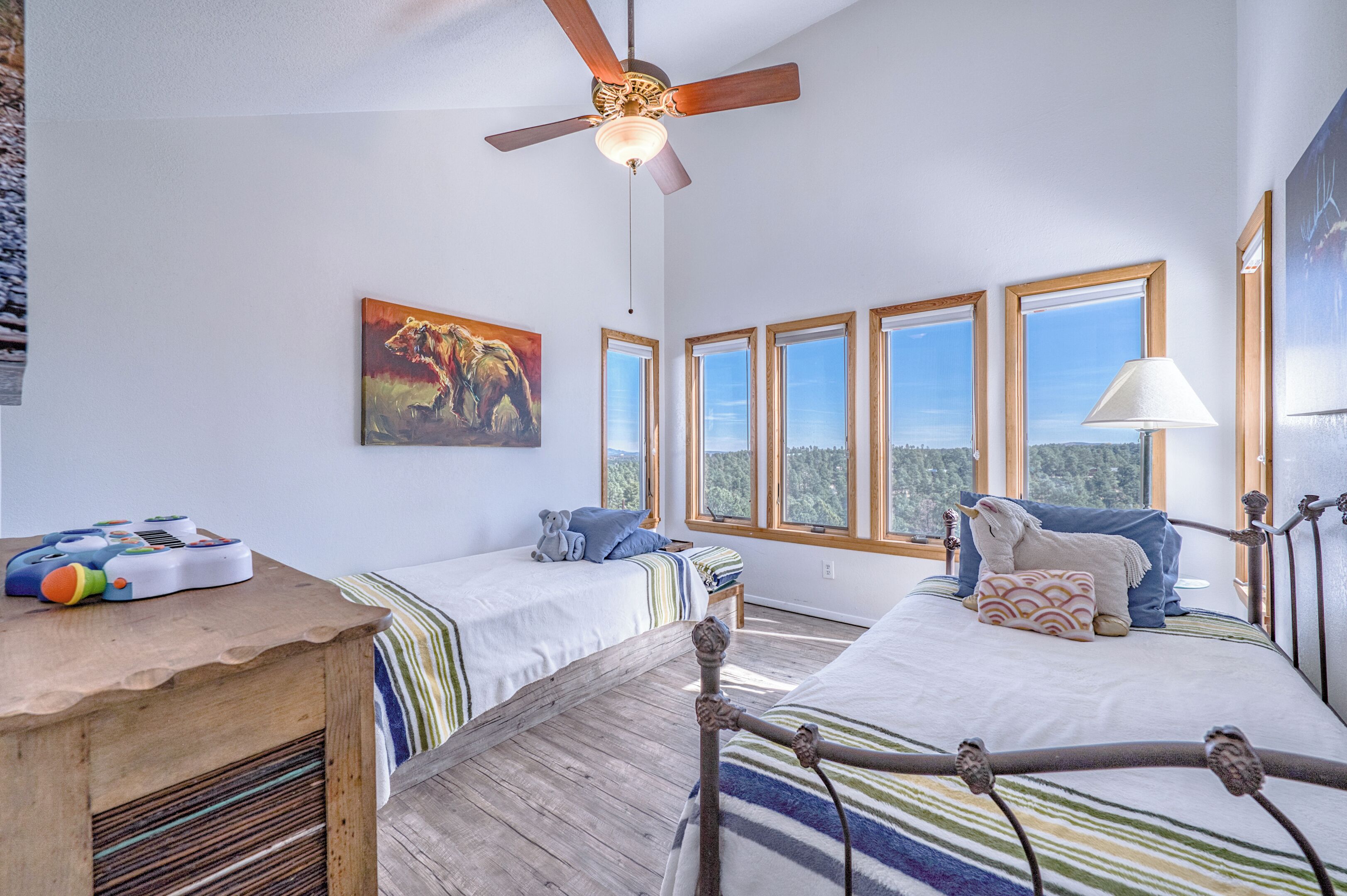 Una habitación con dos camas, ventilador de techo, un cuadro y ventanas grandes.