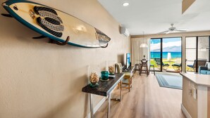 Ein modernes Interieur mit einem an der Wand befestigten Surfbrett, einem Essbereich mit Stühlen und einem Tisch sowie einem Blick auf den Ozean durch große Fenster.
