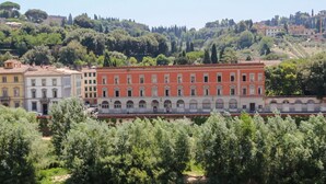 Un bâtiment historique doté d’arcs en plein cintre et d’une façade rouge, entouré d’une végétation luxuriante et bordé par une rivière.
