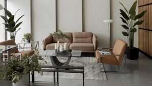 Un salon moderne avec un canapé beige, une table basse en verre et une petite table d’appoint agrémentée d’objets décoratifs. On y trouve deux fauteuils, une lampe sur pied et de grandes plantes en pot.