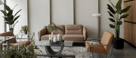 A modern living room with a beige sofa, glass coffee table, and a small side table with decorative items. There are two chairs, a floor lamp, and large potted plants.