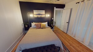 Une chambre à coucher avec un lit, des tables de chevet, une commode, un tableau, une porte et une fenêtre.