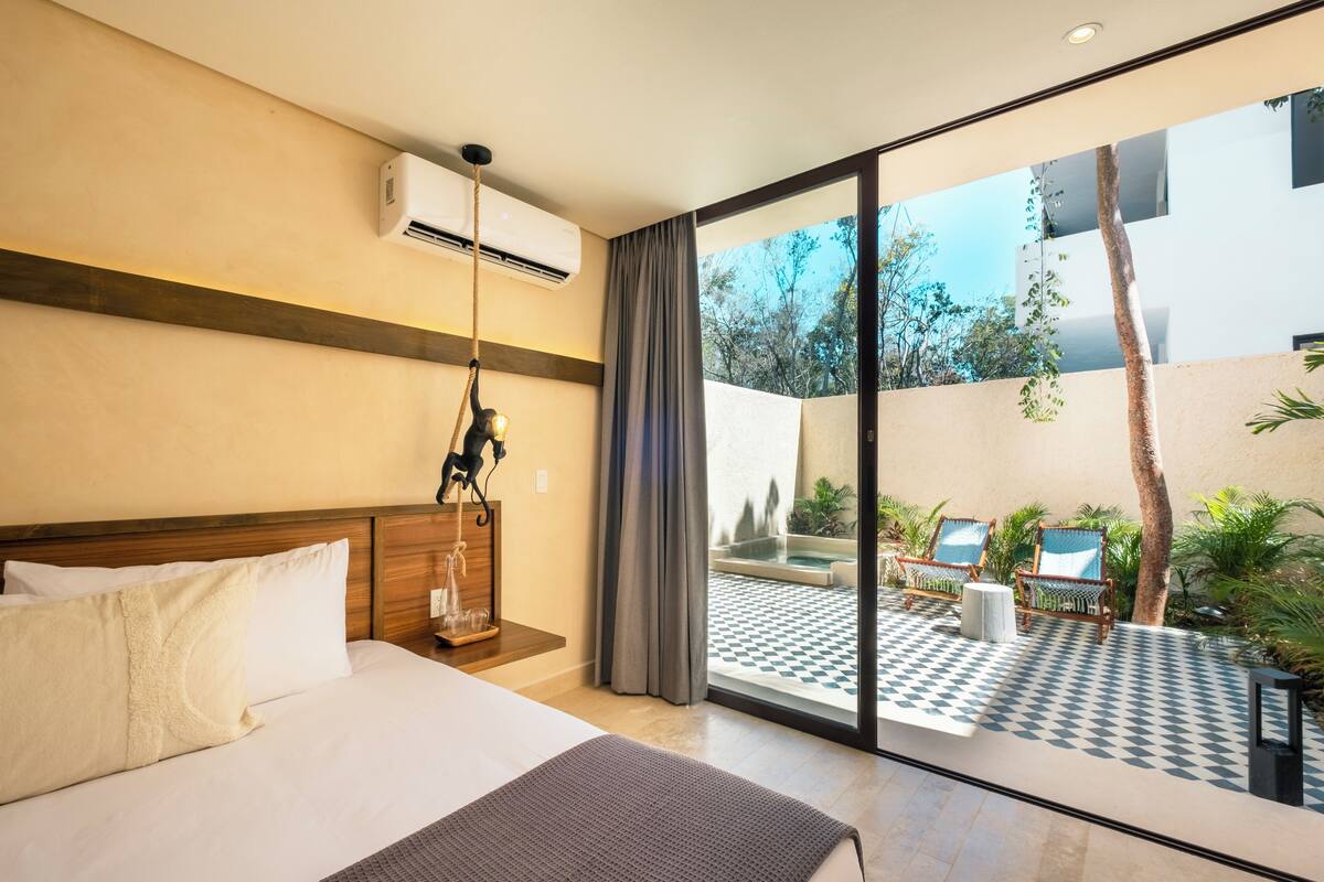 A hotel room with a bed, a large window, and a view of an outdoor area with chairs and a pool.