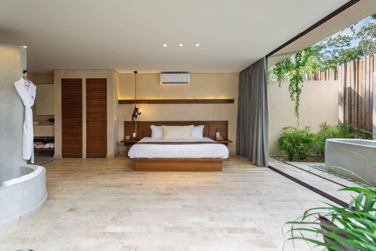 A modern bedroom with a large bed, a built-in bench, a walk-in closet, and a view of greenery outside.