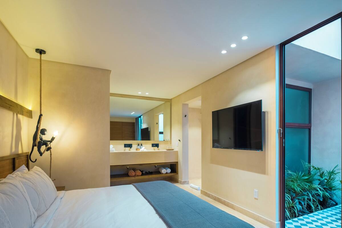 A modern bedroom with a large bed, a TV mounted on the wall, a bathroom with a double sink, and a balcony with plants.