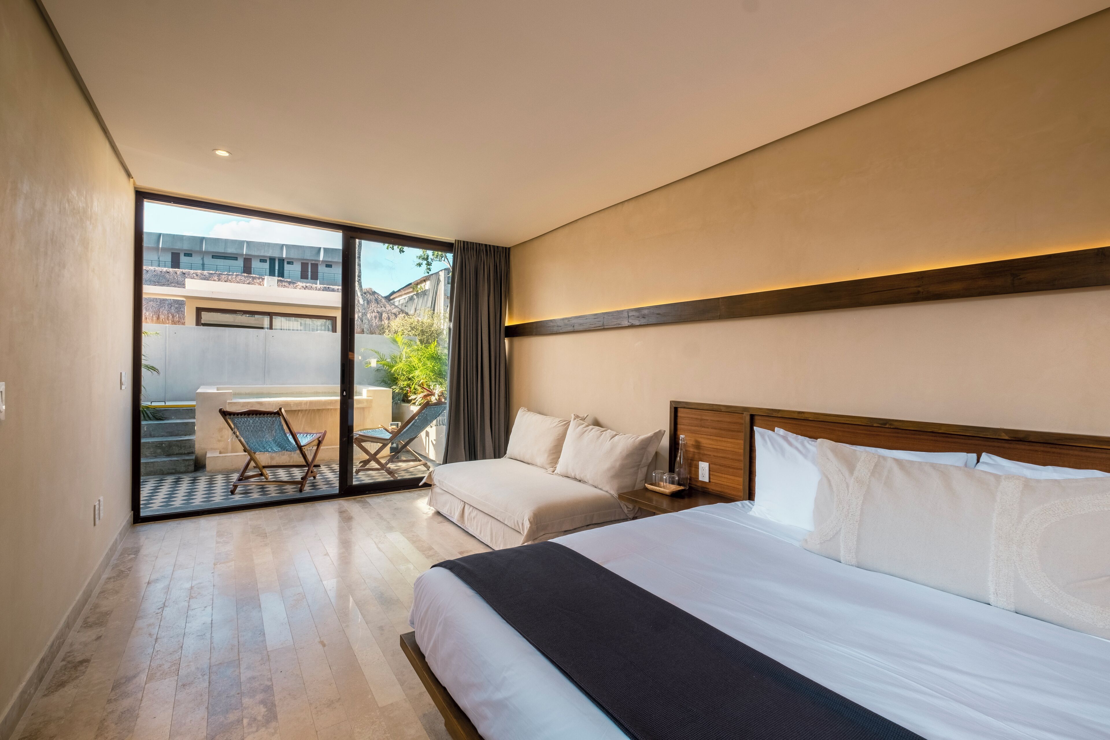 A hotel room with a large bed, a separate sofa, a balcony with a chair, and a view of the outside building.