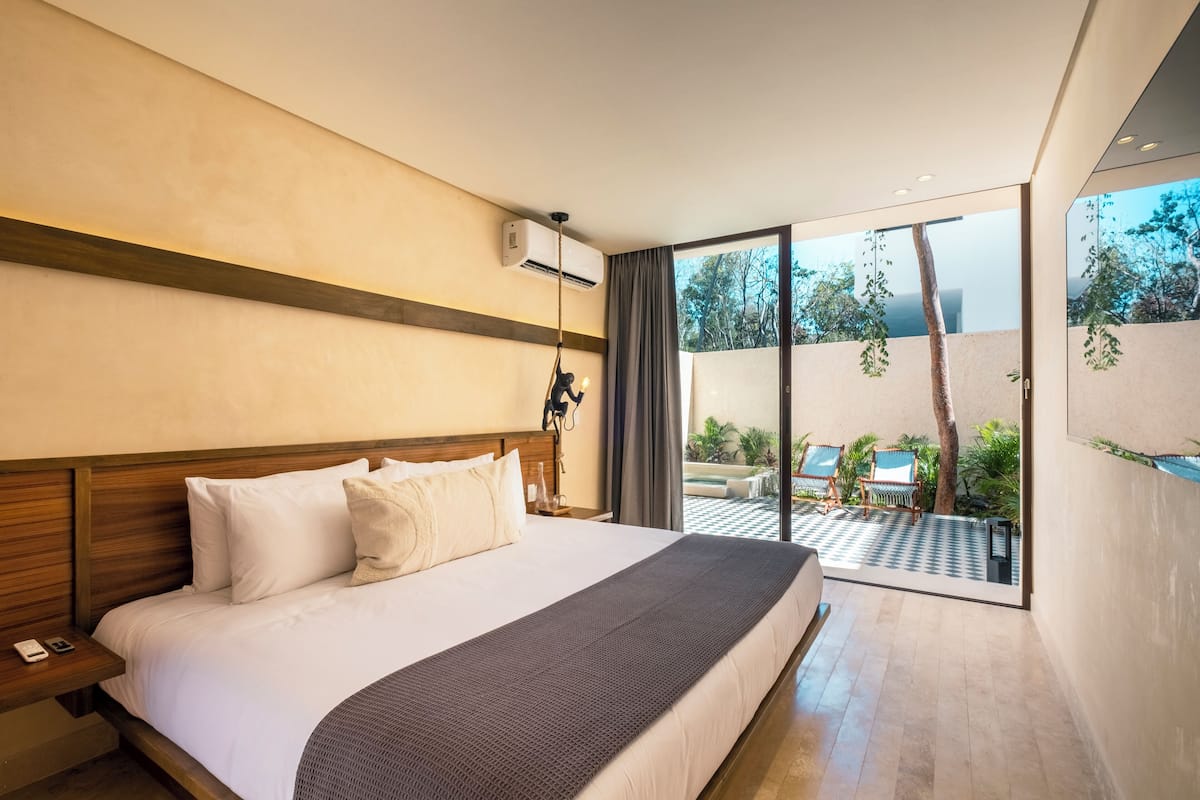 A modern hotel room with a large bed, a wooden headboard, a nightstand, and a view of the outdoor area through large sliding glass doors.