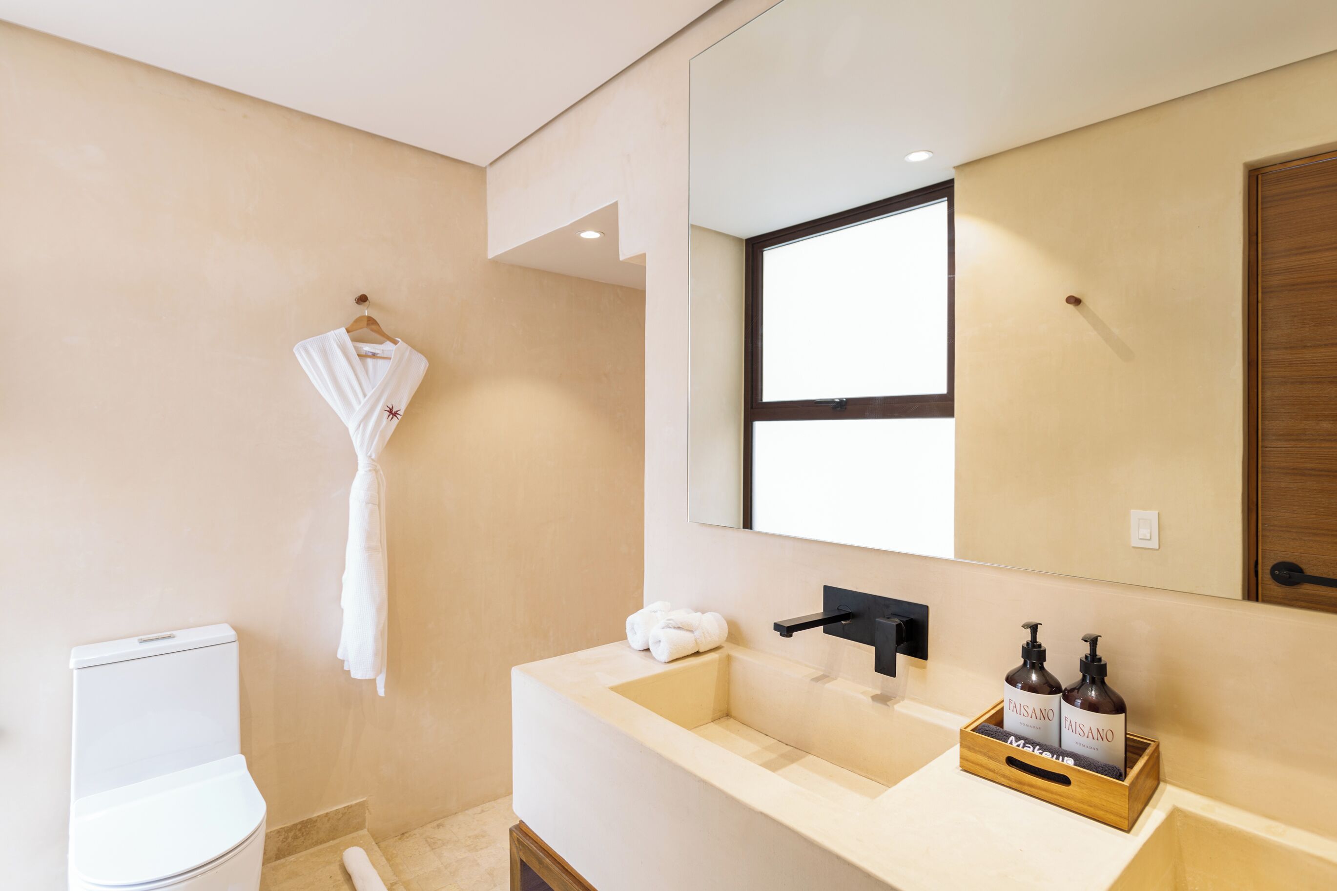 A modern bathroom with a beige wall, a white toilet, a sink with a mirror above it, and a hanging towel.