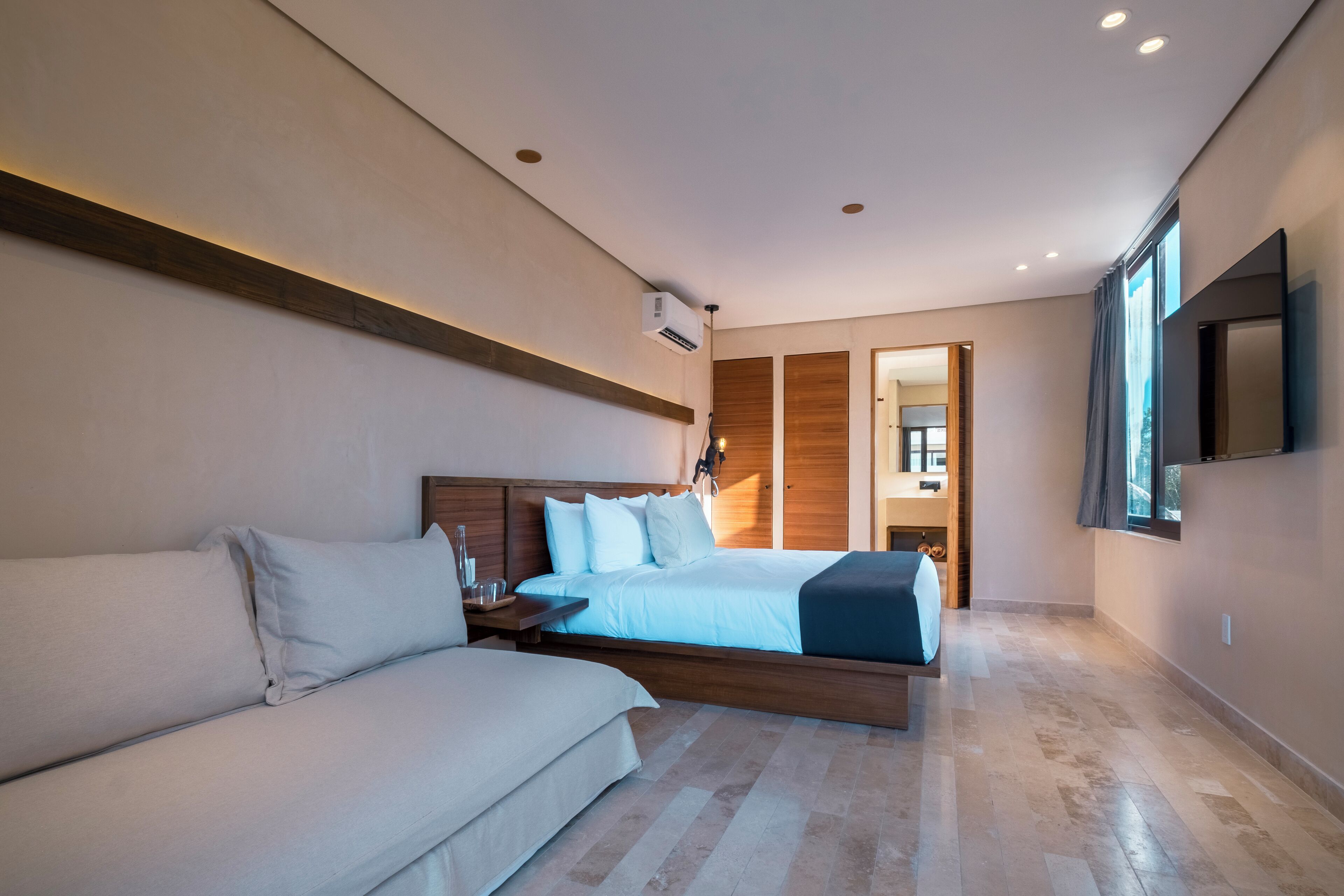 A modern hotel room with a large bed, a sofa, a TV, and a wooden ceiling beam.