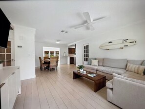 A spacious living area with a ceiling fan, a surfboard decoration, a sofa, a coffee table, and a dining area with chairs and a table.