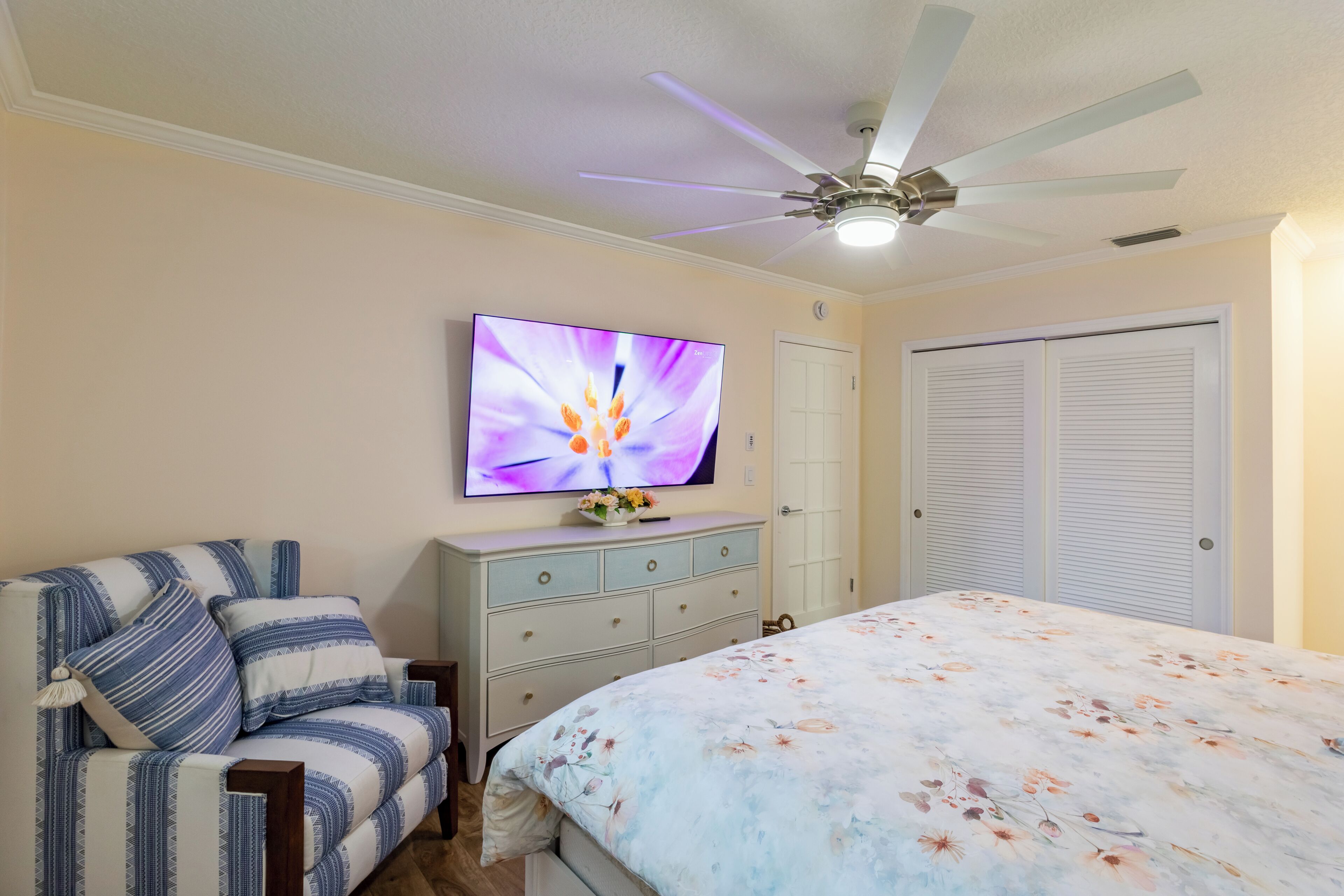 A bedroom with a bed, a television mounted on the wall, a ceiling fan, and a wardrobe.