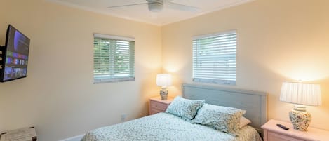 A bedroom with a bed, two bedside tables with lamps, a ceiling fan, and two windows with blinds.
