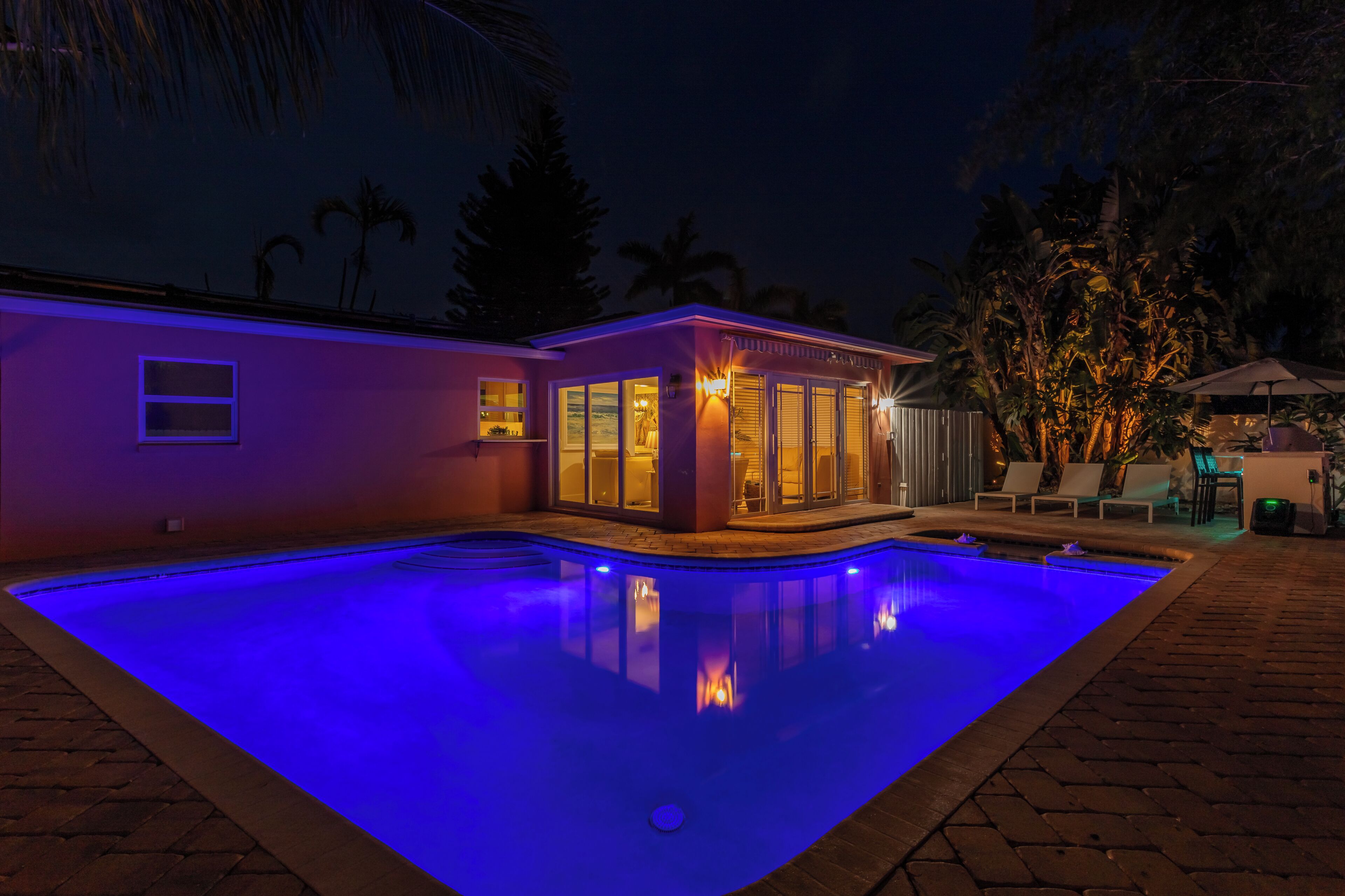 A swimming pool with a blue-lit edge, a small building with glass doors, and a patio area with chairs and a table.