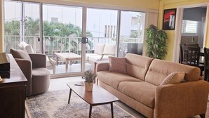 A living room with a sofa, armchairs, a coffee table, and a view through large sliding glass doors to a balcony with outdoor furniture. - Scandia 203 - Sleeps 6, Full Kitchen, Free Parking, and 1 Block to the Beach (Honolulu)