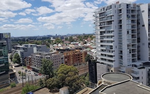 Un edificio de gran altura con varios balcones, vista a un paisaje urbano y una vía ferroviaria en primer plano.