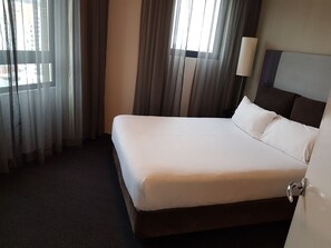 Una habitación de hotel con una cama grande, una lámpara de noche y vistas a la ciudad a través de cortinas translúcidas.