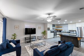 A modern living room with a sofa, coffee table, and TV. A kitchen with a central island and appliances is visible in the background. - Remodeled 4 bedroom 2 bath with sauna home (McAllen)