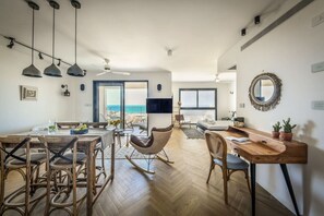 A spacious living area with a dining table, chairs, a rocking chair, a wooden desk, and a large window offering a view.