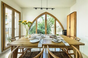A dining table with chairs, a vase with flowers, and a view of greenery through large windows.