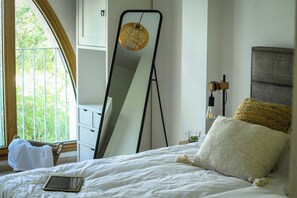 Une chambre à coucher comprenant un lit, une table de chevet, un miroir, une fenêtre donnant sur un paysage verdoyant et une commode blanche.