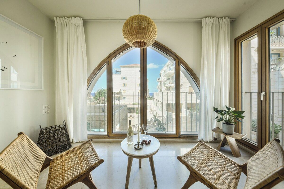 A room with a large arched window, wicker furniture, a small round table with a bottle and glasses, and a hanging light fixture.
