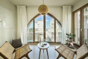 A room with a large arched window, wicker furniture, a small round table with a bottle and glasses, and a hanging light fixture.