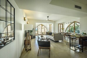A spacious living area with a sofa, a dining table, and a chair. There is a large window providing natural light and a view of the outdoors.