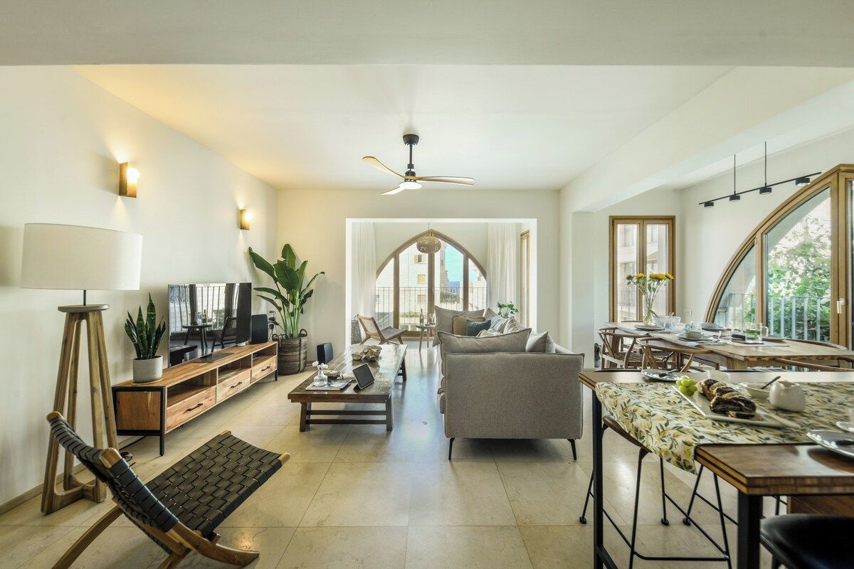 A spacious living area with a sofa, coffee table, and a dining table with chairs. There is a television, a plant, and a ceiling fan.