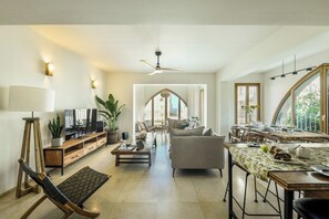 Un grand espace de vie comprenant un canapé, une table basse et une table à manger avec des chaises. On y trouve également un téléviseur, une plante et un ventilateur de plafond.