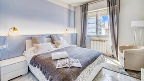 A modern bedroom with a large bed, bedside lamps, a reading light, a window with curtains, and a view of buildings. - Bedo House - Beachfront Sea View Apartment in Follonica, Follonica, Italy (Follonica)