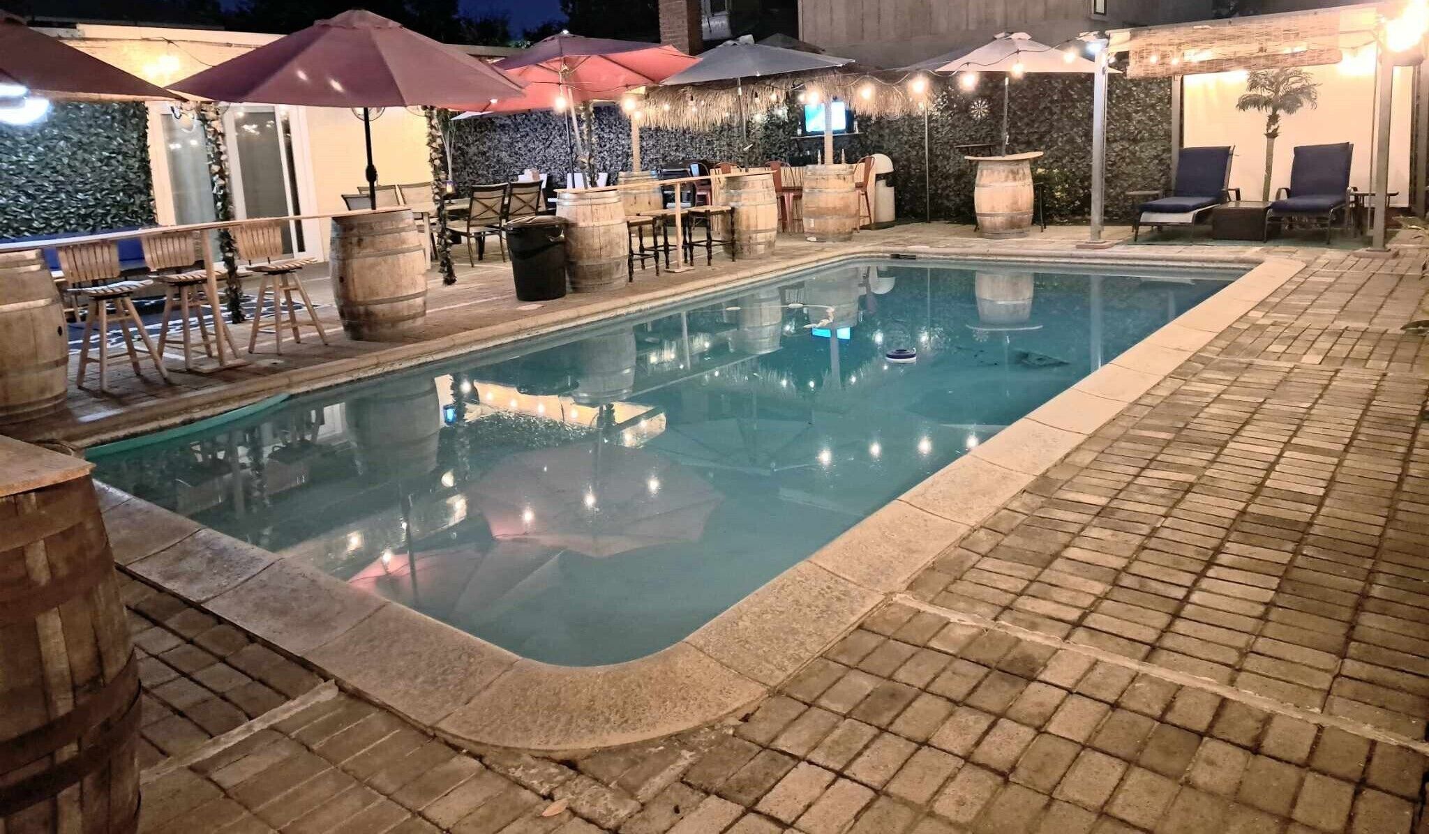 An outdoor swimming pool with wooden barrels and lounge chairs, surrounded by umbrellas and stone walls.