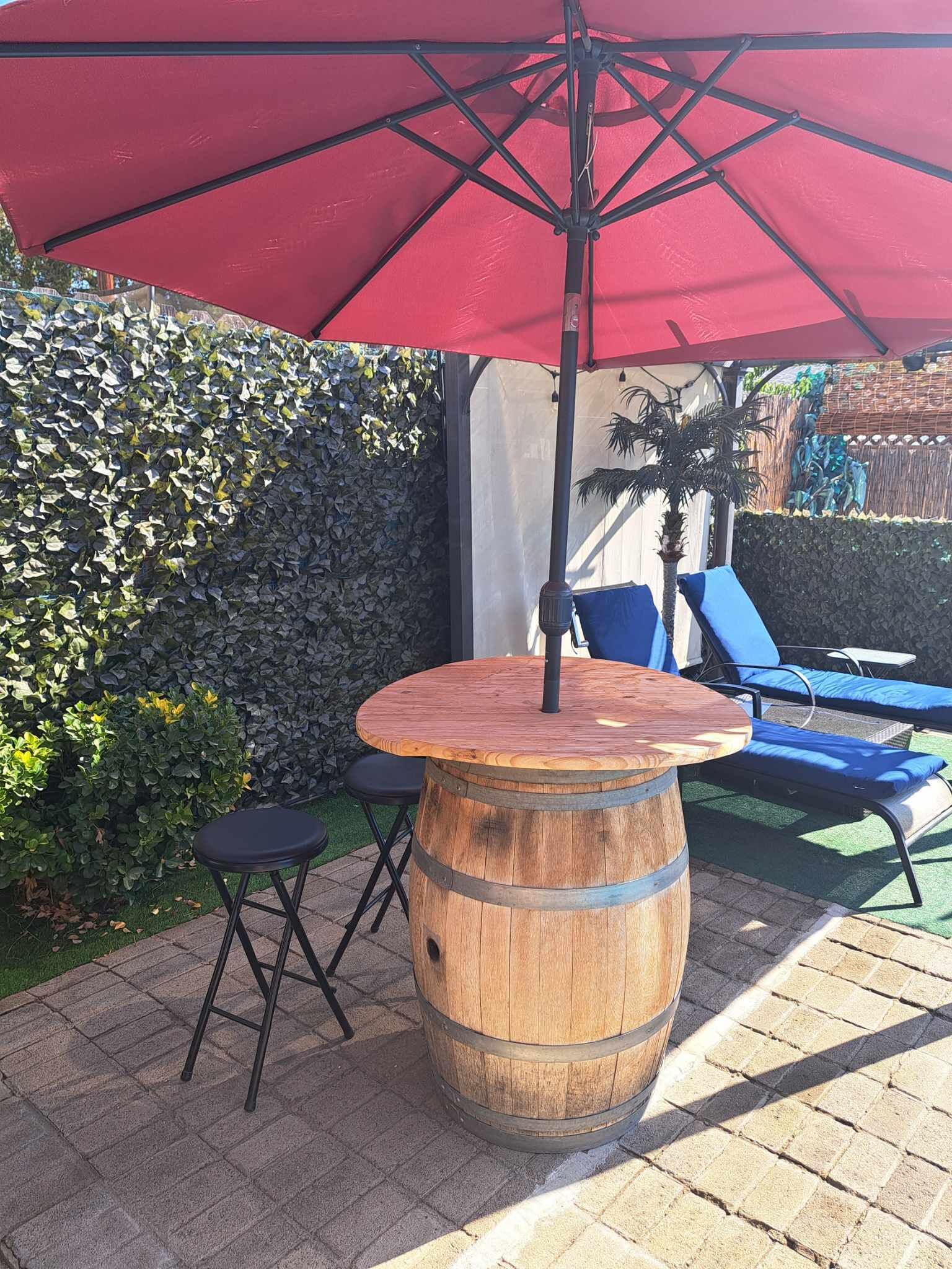 A wooden barrel table with a red umbrella, two black chairs, and blue lounge chairs in a garden setting.