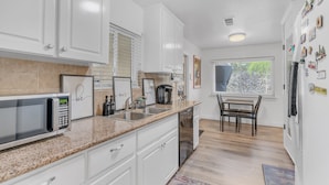 A modern kitchen with white cabinets, a granite countertop, a built-in microwave, a coffee maker, and a dining area with a table and chairs. - Tropical Paradise  (Fresno)