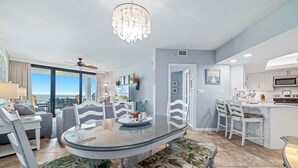 A modern dining area with a glass table, white chairs, and a bench with a leafy pattern. - Summer House 705B | Gorgeous Beach Front, Huge Deck, Great Location (Orange Beach)
