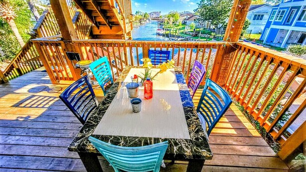 Une terrasse en bois avec une table et des chaises, donnant sur un plan d’eau et des bâtiments.