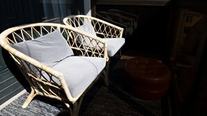 Deux fauteuils en osier avec des coussins gris, un tapis tissé foncé et un pouf rouge dans une pièce dotée d’une fenêtre et d’un mur sombre.