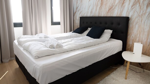 Un lit bien fait, avec du linge de lit blanc et des serviettes pliées, une tête de lit noire et une petite table de chevet blanche avec une lampe.