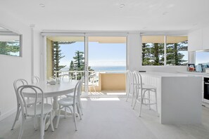 Een moderne keuken met witte kasten en een groot eiland, een eethoek met witte stoelen, en een balkon met uitzicht op zee en bomen.