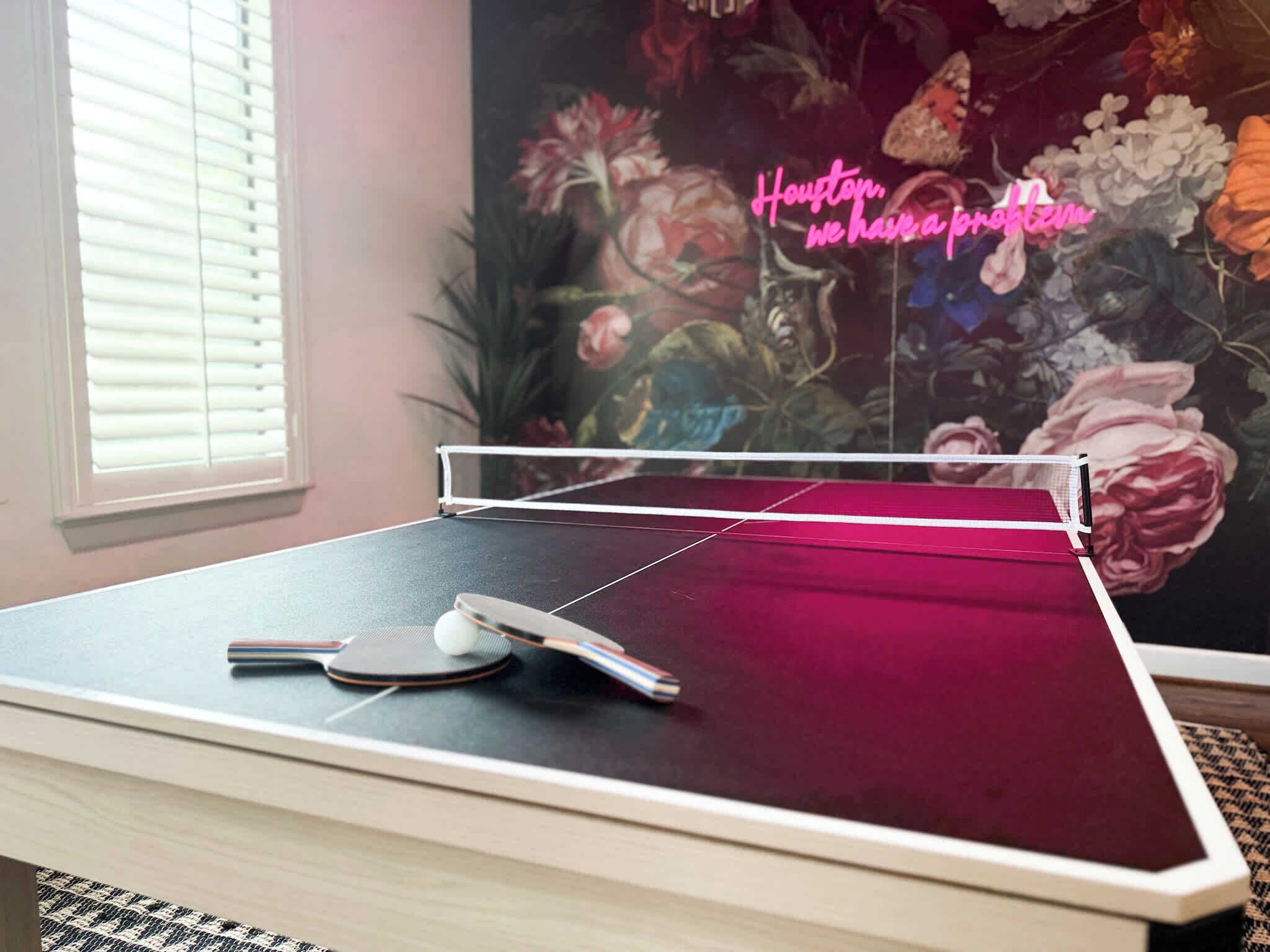 A ping pong table in a room with a floral mural on the wall and a window with blinds.