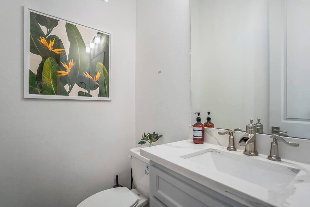 A bathroom with a white sink, two faucets, a mirror, and a toilet. There is a framed picture of flowers on the wall.