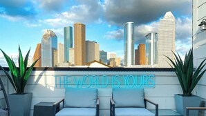An outdoor seating area with two chairs and a fire feature, a neon sign stating "THE WORLD IS YOURS", and a city skyline in the background. - Urban Jungle Retreat in the HTX Skyline (Houston)