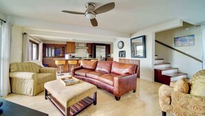 Un salon avec un canapé en cuir marron, un fauteuil à rayures et une table basse en bois. On y trouve un ventilateur de plafond, une cuisine à l’arrière-plan et un escalier menant à l’étage supérieur.