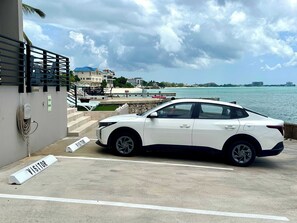 A white car parked in a visitor parking area with a view of the sea and buildings in the background. - Seven Mile View #3A - Oceanfront Studio & Pool (West Bay)