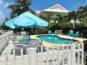 A swimming pool with a white fence, blue chairs, and a blue umbrella.